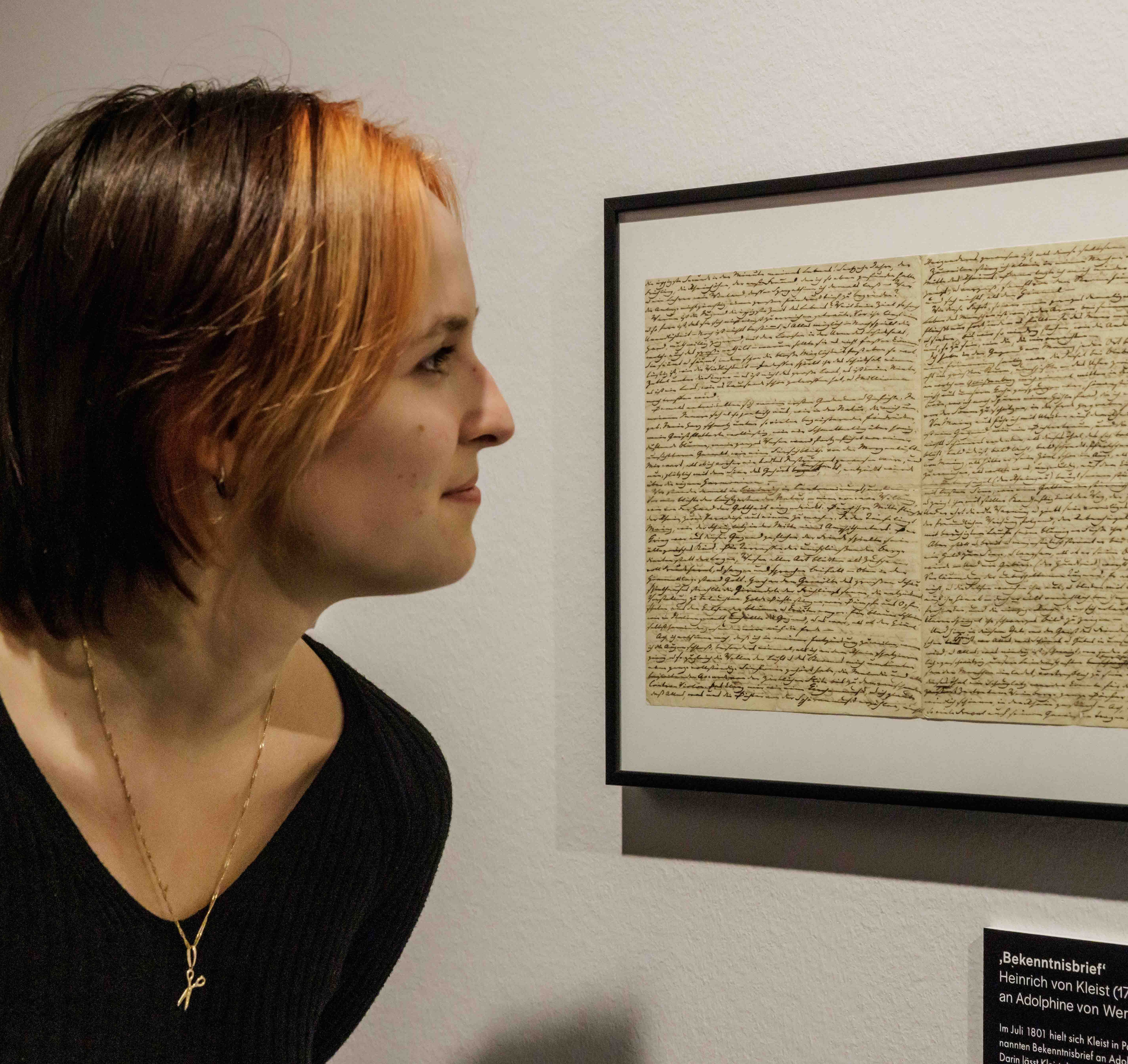 Osoba o krótkich włosach i ubrana w czarny sweter ogląda oprawiony, ręcznie napisany list na ścianie. Miejsce akcji to muzeum lub galeria, co budzi ciekawość.