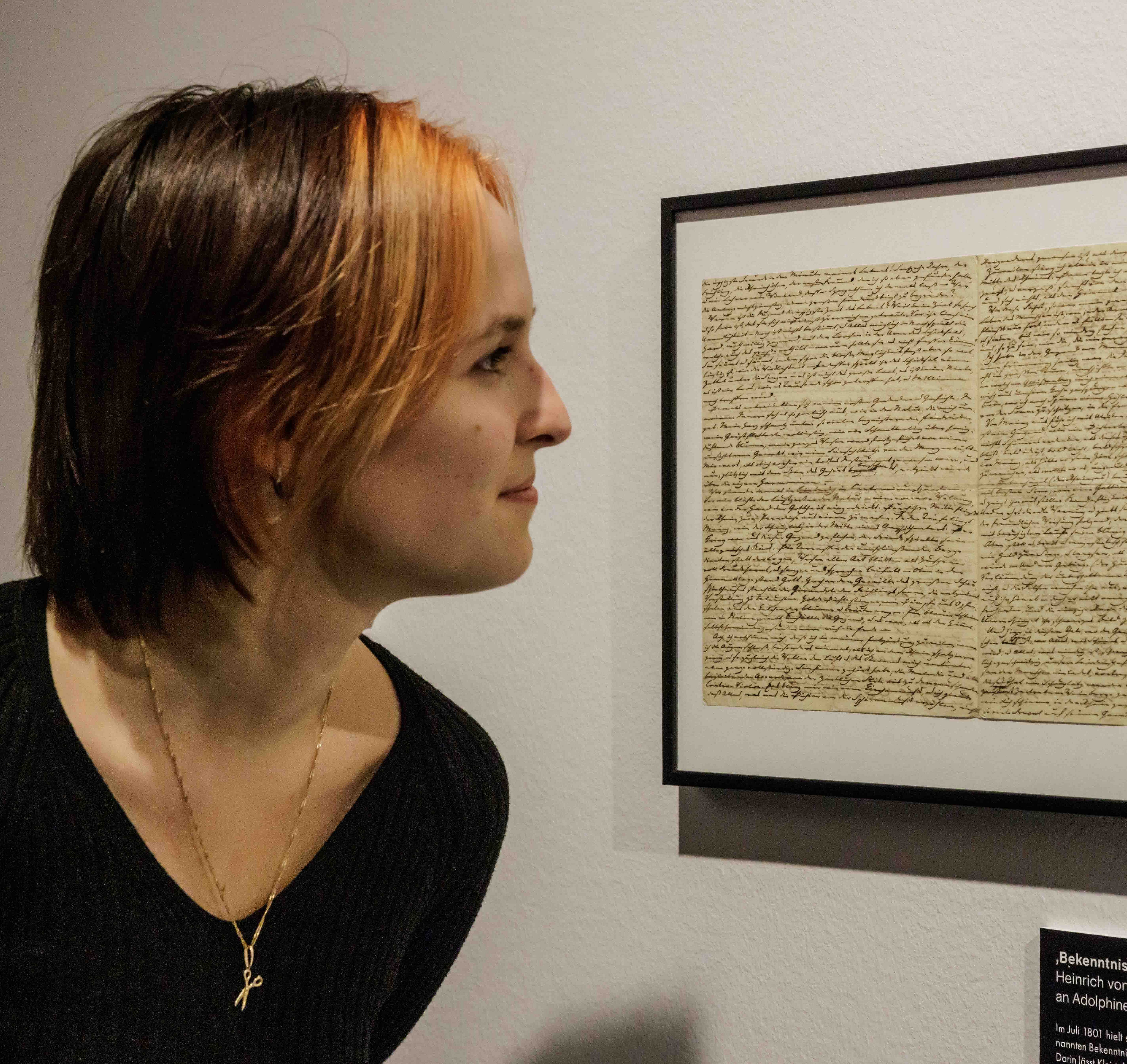 Eine Person mit kurzen Haaren und einem schwarzen Pullover betrachtet einen gerahmten, handgeschriebenen Brief an einer Wand. Der Schauplatz ist ein Museum oder eine Galerie, was Neugierde vermittelt.