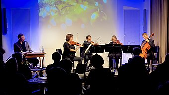 Vier Musikerinnen und Musiker spielen und eine Schauspieler liest auf einer Bühne vor Publikum, im Hintergrund ist eine festliche Projektion mit Lichterpunkten und Kugeln zu sehen.