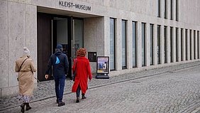 Drei Personen gehen auf den Eingang eines modernen Gebäudes mit heller Steinfassade zu. Über der Tür steht in klaren Großbuchstaben KLEIST-MUSEUM. Links neben dem Eingang steht ein Aufsteller mit Plakat. Das Kopfsteinpflaster vor dem Gebäude ist sauber und gleichmäßig verlegt.