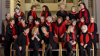 Gruppe von Frauen und einem Mann in schwarzer Kleidung mit roten Schals sitzt und steht in mehreren Reihen in einem hellen Innenraum.