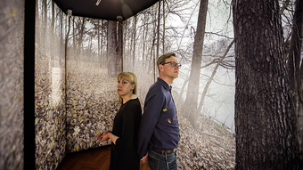 Zwei Personen stehen Rücken an Rücken in einem Ausstellungsraum. Die Wände sind vollständig mit großformatigen Fotografien eines herbstlichen Waldbodens und kahler Bäume bedeckt. Die Decke ist dunkel, eine einzelne Lampe leuchtet von oben. Die Personen blicken in entgegengesetzte Richtungen, eine trägt ein schwarzes Oberteil, die andere ein dunkelblaues. Der Raum wirkt ruhig und nachdenklich.