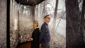 Zwei Personen stehen Rücken an Rücken in einem Ausstellungsraum. Die Wände sind vollständig mit großformatigen Fotografien eines herbstlichen Waldbodens und kahler Bäume bedeckt. Die Decke ist dunkel, eine einzelne Lampe leuchtet von oben. Die Personen blicken in entgegengesetzte Richtungen, eine trägt ein schwarzes Oberteil, die andere ein dunkelblaues. Der Raum wirkt ruhig und nachdenklich.