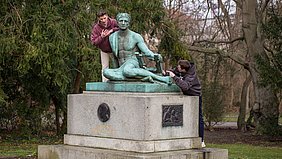 Eine bronzene Statue auf einem steinernen Sockel steht in einer Parkanlage. Die Figur stellt eine sitzende Person mit einer antiken Leier dar. Zwei Personen lehnen sich von beiden Seiten an die Statue, eine links, eine rechts. Beide Personen tragen Freizeitkleidung. Im Hintergrund sind Bäume und Sträucher ohne Laub zu sehen.