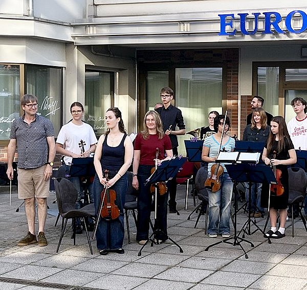 Eine Gruppe von Menschen steht mit Streichinstrumenten und Notenständern im Halbkreis vor einem Gebäude. Die meisten halten Geigen, Bratschen oder Celli, einige tragen lässige Kleidung, andere dunkle Oberteile. Im Hintergrund ist ein Gebäudeeingang mit großen Fenstern und Glastüren zu sehen. Über dem Eingang befindet sich ein blaues Schild mit dem Schriftzug ‚EUROPA-UNIVE'RSITÄT‘. Die Stimmung wirkt konzentriert und freundlich.