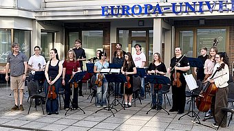 Eine Gruppe von Menschen steht mit Streichinstrumenten und Notenständern im Halbkreis vor einem Gebäude. Die meisten halten Geigen, Bratschen oder Celli, einige tragen lässige Kleidung, andere dunkle Oberteile. Im Hintergrund ist ein Gebäudeeingang mit großen Fenstern und Glastüren zu sehen. Über dem Eingang befindet sich ein blaues Schild mit dem Schriftzug ‚EUROPA-UNIVE'RSITÄT‘. Die Stimmung wirkt konzentriert und freundlich.