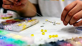 Zwei Hände fädeln kleine, bunte Perlen auf einen dünnen Faden. Im Vordergrund liegen mehrere Sortierboxen mit unterschiedlich farbigen Perlen, darunter auch gelbe Perlen mit aufgemalten Gesichtern. Die Person trägt ein helles T-Shirt. Der Fokus liegt auf den Händen und den Perlen, der Hintergrund ist unscharf.