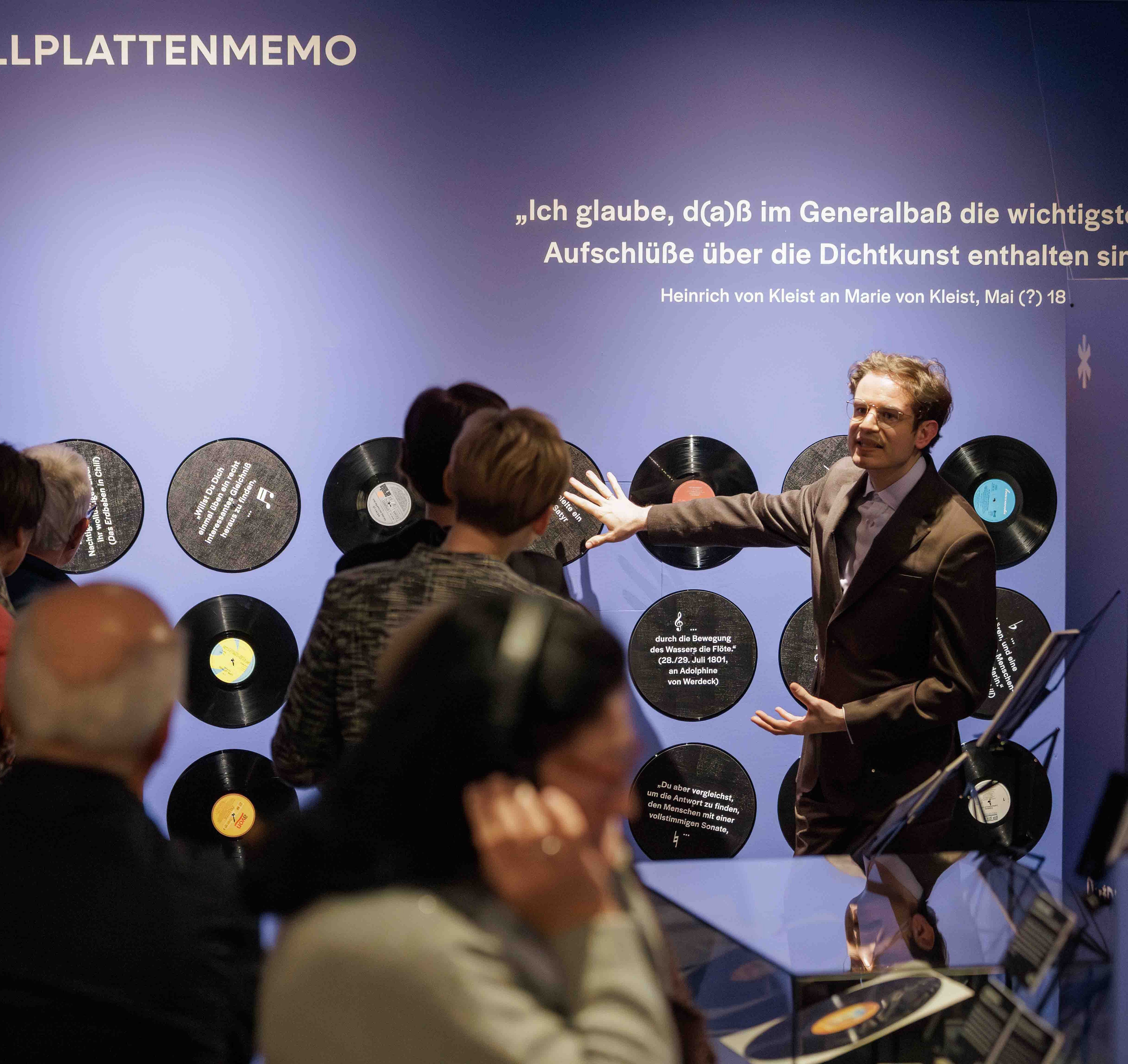 Ein Mann im Anzug steht vor einer blauen Wand mit Schallplatten und spricht zu einer Gruppe von Menschen. Die Schallplatten sind in einer Reihe an der Wand angebracht. Über den Schallplatten steht in großen weißen Buchstaben ein Zitat von Heinrich von Kleist. Die Zuhörerinnen und Zuhörer stehen dicht beieinander und blicken aufmerksam auf den Mann, der mit einer ausladenden Geste spricht. Die Szene spielt in einem Ausstellungsraum mit gedämpfter Beleuchtung.