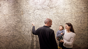 Eine großformatige, detaillierte Landkarte mit zahlreichen Städtenamen und Grenzlinien bedeckt die gesamte Wand. Im Vordergrund stehen drei Personen: Eine erwachsene Person mit kurzem Haar und dunkler Kleidung zeigt mit ausgestrecktem Arm auf einen Bereich der Karte. Eine weitere erwachsene Person mit langen Haaren hält ein Kind auf dem Arm. Alle blicken in Richtung der Karte. Die Szene ist hell ausgeleuchtet.