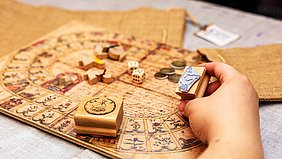 Hand hält einen Holzspielstein mit Gänsemotiv über einem historischen Spielbrett mit Würfeln und weiteren Spielsteinen