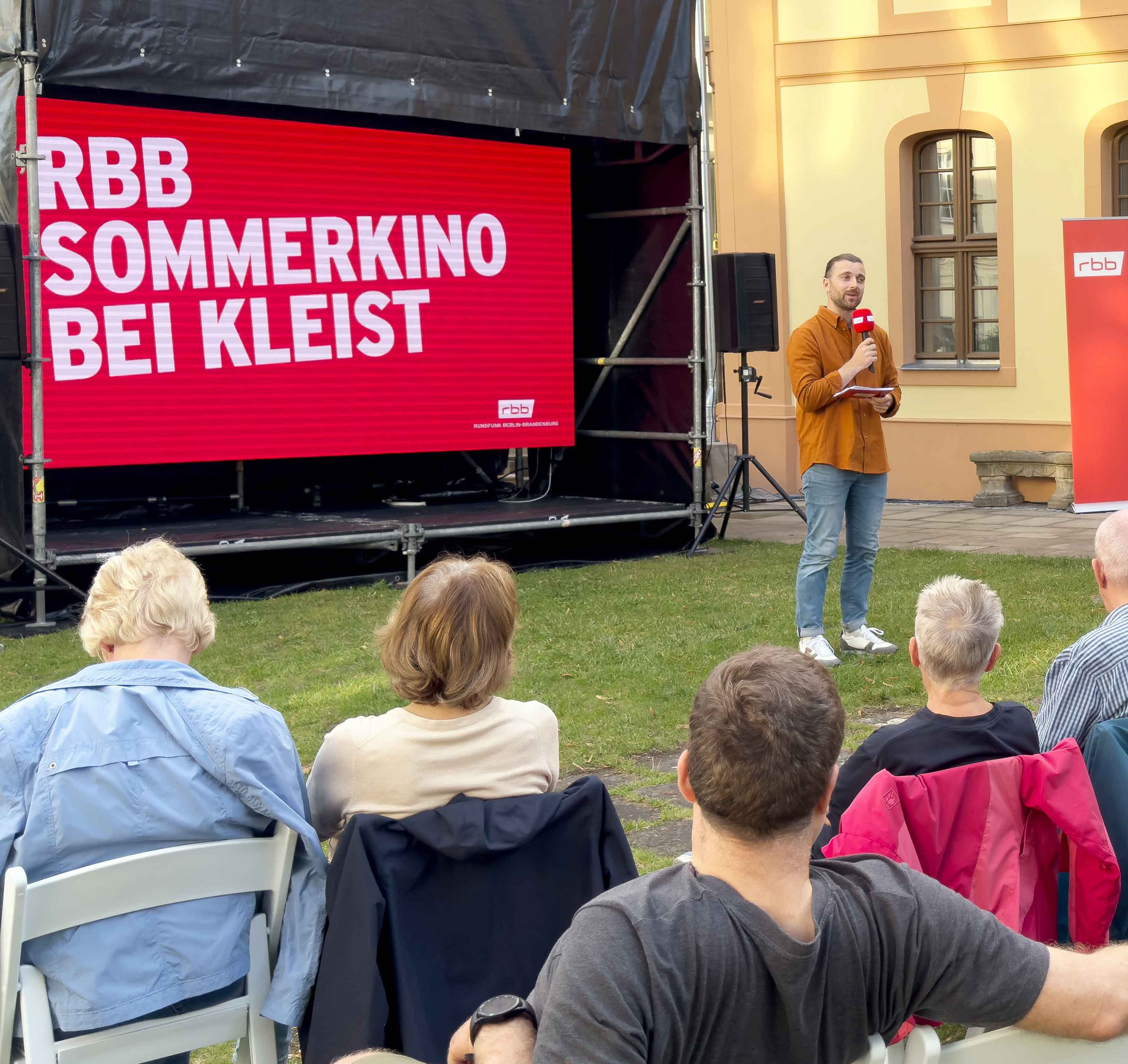  Im Innenhof eines historischen Gebäudes sitzen Menschen auf weißen Klappstühlen und blicken auf eine große Leinwand mit rotem Hintergrund. Darauf steht in weißen Großbuchstaben: ‚RBB SOMMERKINO BEI KLEIST‘. Ein Mann mit Mikrofon steht vor der Leinwand und spricht zum Publikum. Im Hintergrund sind gelb gestrichene Fassaden mit großen Fenstern und ein Türbogen zu sehen. Die Szene spielt bei Tageslicht auf einer Rasenfläche.