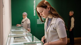 Junge Frau betrachtet ausgestellte Dokumente in einer Vitrine vor einer grünen Wand mit dem Schriftzug ‚SCHRIFTSTELLERLEBEN‘
