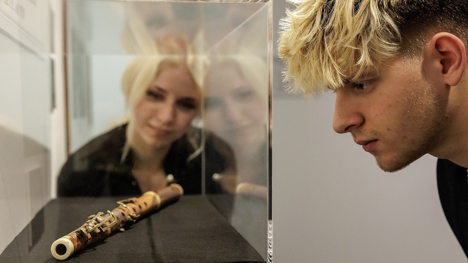 Person looking at a historical wooden flute with metal keys which rests on a black cloth inside a clear display case. The flute has a cylindrical shape and visible key mechanisms along its body. The background is plain and neutral, with a glass wall reflecting the flute and the surrounding area.