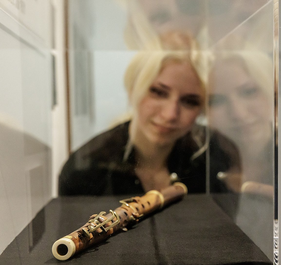 Wgląd w wystawę specjalną ©Gordon Welters Osoba patrząca na zabytkowy drewniany flet z metalowymi klapami, który spoczywa na czarnym suknem wewnątrz przezroczystej gabloty. Flet ma cylindryczny kształt i widoczne mechanizmy klap wzdłuż korpusu. Tło jest proste i neutralne, a szklana ściana odbija flet i otaczającą go przestrzeń.
