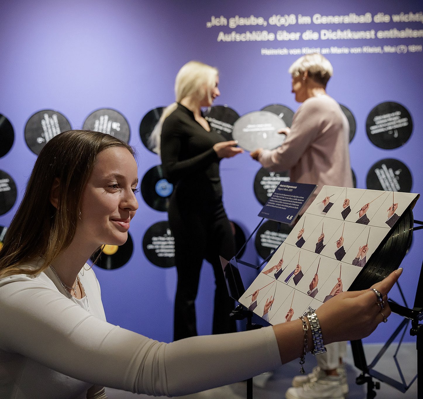 Against a blue wall decorated with vinyl record motifs and a white quote, a person in the foreground holds a square board with a graphic design depicting hands in various gestures. In the background, there are two people, one of whom is holding a vinyl record. The scene takes place at a music or art exhibition.