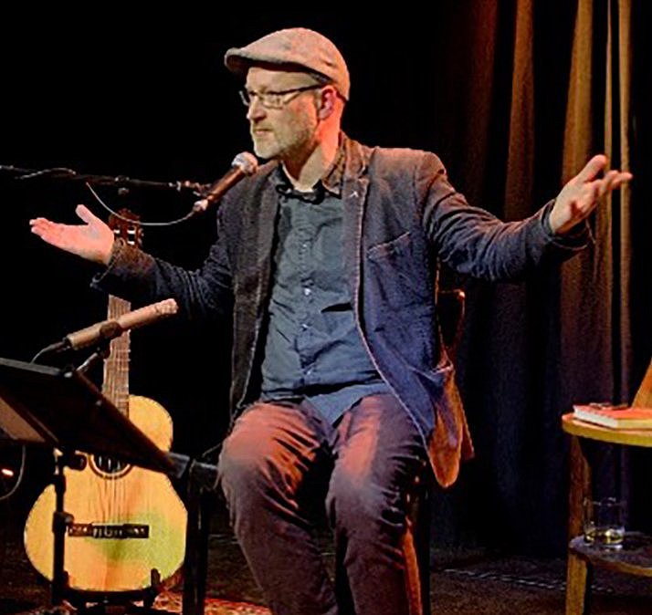 Ein Musiker sitzt auf einer Bühne, spricht mit gestikulierenden Händen und hat eine Gitarre sowie ein Bild auf einem Tisch neben sich.