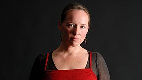 Eine Person mit mittellangem, zurückgebundenem Haar trägt ein rotes Oberteil mit schmalen Trägern über einem schwarzen, langärmeligen Shirt. Der Hintergrund ist einfarbig dunkel gehalten.