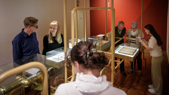 Mehrere Personen stehen in einem Ausstellungsraum mit rötlich gestrichener Wand. Sie betrachten Exponate in verglasten Vitrinen, die auf goldfarbenen Metallgestellen ruhen. Der Boden besteht aus Holzparkett. Die Beleuchtung ist dezent und schafft eine ruhige Atmosphäre.