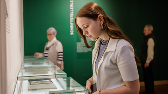 Eine junge Frau mit langen, rötlichen Haaren betrachtet aufmerksam ein Schriftstück in einer Vitrine. Sie trägt eine helle Strickjacke über einem hellblauen Oberteil und silberne Ohrringe. Im Hintergrund stehen zwei weitere Personen, eine Frau mit kurzen grauen Haaren und ein Mann mit grauem Haar, die ebenfalls die Ausstellung betrachten. Die Wände sind dunkelgrün gestrichen und mit weißer Schrift versehen. Die Vitrinen enthalten verschiedene Dokumente und Schriftstücke, die flach ausgelegt sind. Der Raum wirkt ruhig und konzentriert.
