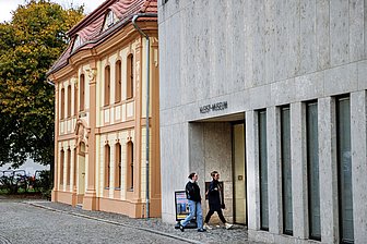 Zwei Frauen gehen nebeneinander am Eingang eines modernen Gebäudes mit der Aufschrift ‚Kleist-Museum‘ vorbei, daneben steht ein historisches Haus.