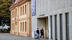Zwei Frauen gehen nebeneinander am Eingang eines modernen Gebäudes mit der Aufschrift ‚Kleist-Museum‘ vorbei, daneben steht ein historisches Haus.