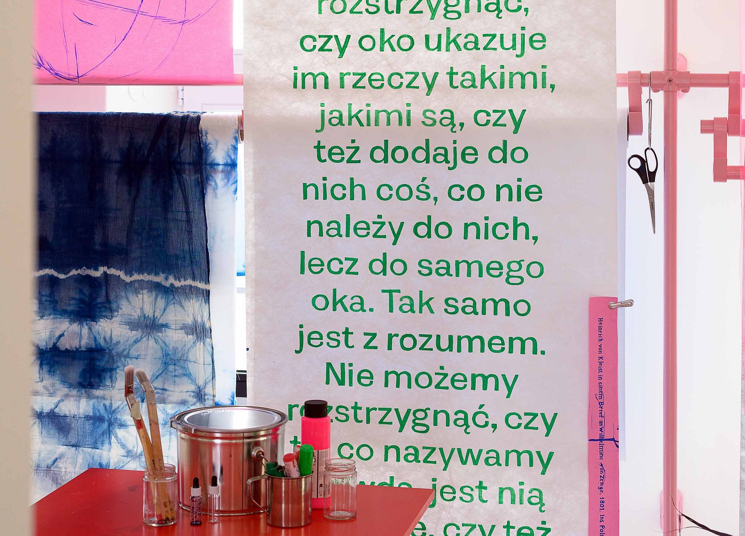 Obok stołu warsztatowego z pędzlami, szklankami i narzędziami wisi wysoka rolka papieru z tekstem w języku polskim.
