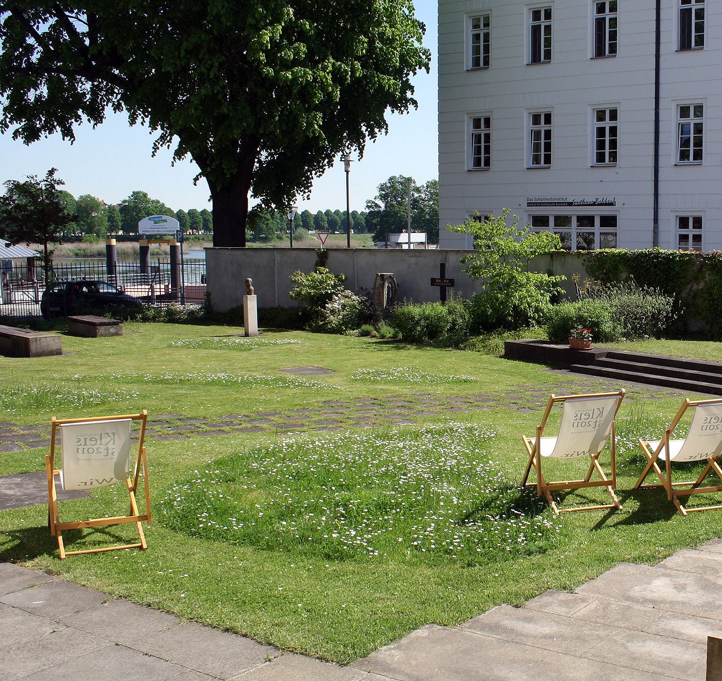 Ein gepflegter Garten mit Rasenfläche, auf der drei Holzliegestühle mit heller Bespannung stehen. Im Vordergrund steht ein Blumentopf mit einer grünen Pflanze auf einer gepflasterten Terrasse. Im Hintergrund befindet sich ein großes, weißes Gebäude mit mehreren Fenstern und einem Eingang mit Vordach. Einzelne Bäume und Sträucher säumen den Garten, und eine niedrige Mauer trennt das Gelände von der Umgebung. Die Szene wirkt ruhig und einladend.