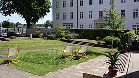 Ein gepflegter Garten mit Rasenfläche, auf der drei Holzliegestühle mit heller Bespannung stehen. Im Vordergrund steht ein Blumentopf mit einer grünen Pflanze auf einer gepflasterten Terrasse. Im Hintergrund befindet sich ein großes, weißes Gebäude mit mehreren Fenstern und einem Eingang mit Vordach. Einzelne Bäume und Sträucher säumen den Garten, und eine niedrige Mauer trennt das Gelände von der Umgebung. Die Szene wirkt ruhig und einladend.