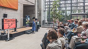 Mehrere Personen sitzen in einem modernen Saal mit Glasfassade und hören zwei stehenden Rednerinnen zu, links steht ein Bildschirm mit der Aufschrift ‚7Sachen‘.