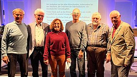 Sechs Erwachsene stehen nebeneinander vor einer Leinwand mit projiziertem Text.