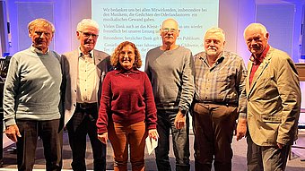 Sechs Erwachsene stehen nebeneinander vor einer Leinwand mit projiziertem Text.