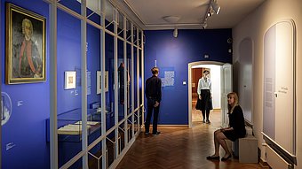 Mehrere Personen in einem Ausstellungsraum mit blauen Wänden, Vitrinen und einem gerahmten Porträt an der Wand.