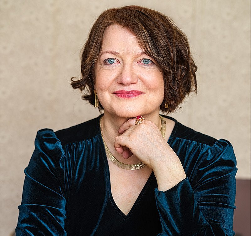 Eleonora Hummel ©get-shot.de Eine Frau mit kurzem, braunem Haar sitzt in einem dunkelblauen Samtkleid mit Puffärmeln und einer Perlenkette. Sie stützt ihren Kopf auf die Hand und blickt nach vorne.